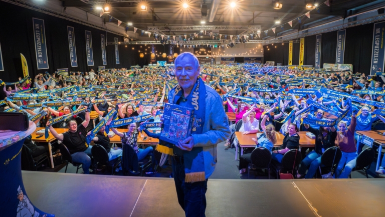 Een man op een podium houdt een puzzel vast terwijl een grote menigte juichend blauwe en gele sjaals omhooghoudt.