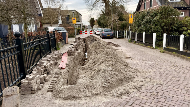 Geopende straat in een woonwijk met een geul gevuld met zand en opgebroken stoeptegels aan de zijkant.