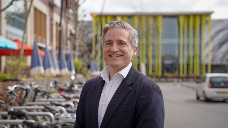 Man in een donkerblauwe blazer glimlacht buiten, met fietsen en een modern gebouw op de achtergrond.