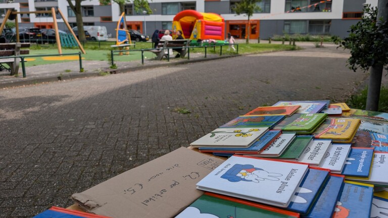 Kinderboeken liggen op een tafel in een speeltuin, met een springkussen en een paar mensen op de achtergrond.