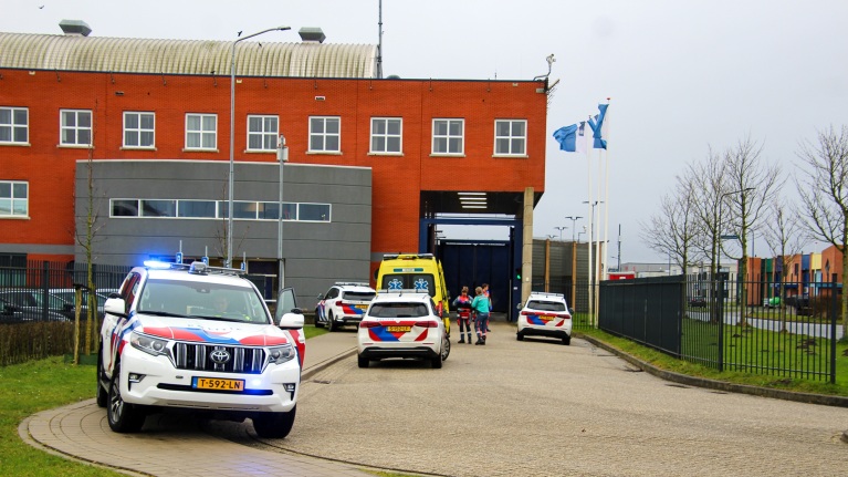 Politieauto's, een ambulance en personeel voor een gebouw met rode bakstenen en vlaggen op een bewolkte dag.