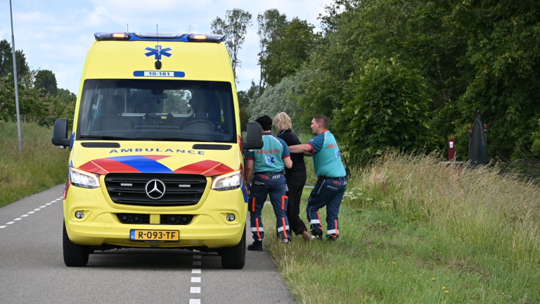 Paramedici helpen een persoon naast een gele ambulance op een landweg, omringd door groen.