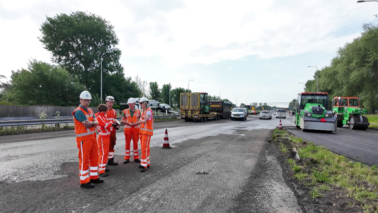 Werknemers in oranje veiligheidskleding en helmen staan op een weg bij een wegenbouwproject, omgeven door machines en auto's.