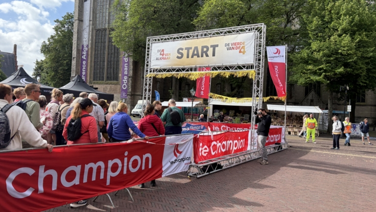 Groep mensen wacht bij de start van een wandeltocht in Alkmaar, met rode Champion-banners en een kerk op de achtergrond.