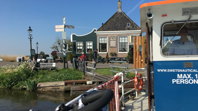 Een veerboot aan de kade met een historisch gebouw en een persoon op een bankje in een schilderachtig dorp.