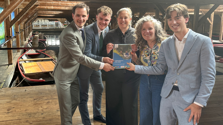 Een groep van vijf mensen poseert met een document genaamd "Coalitieakkoord 2026-2030" in een houten gebouw met boten op de achtergrond.