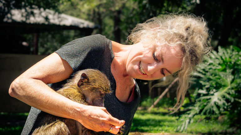 Vrouw met blonde haren glimlacht terwijl ze een kleine aap vasthoudt in een groene, zonnige omgeving.
