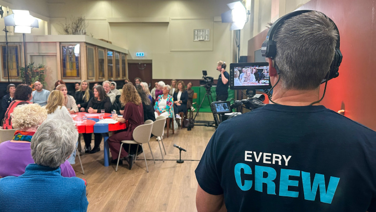 Een cameraman filmt een talkshow met een groep mensen rond een tafel en publiek op de achtergrond.