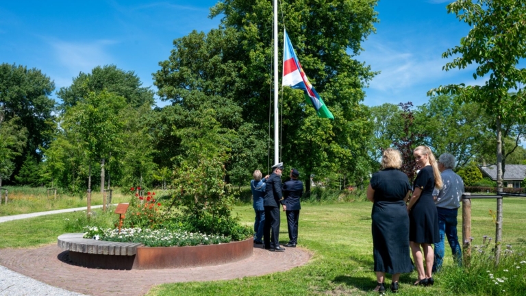 Mensen hijsen een vlag in een parkachtige omgeving, omgeven door bomen en een bloembed.
