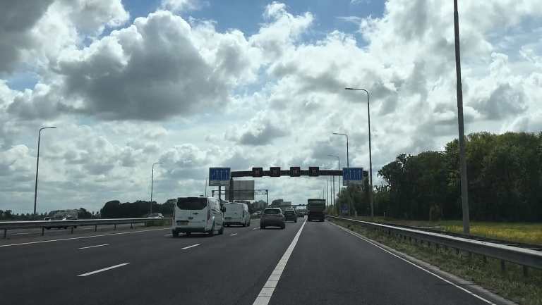 Een snelweg met auto's die onder een portaal met snelheidsbeperkingen van 90 km/u rijden, omgeven door wolkenluchten.
