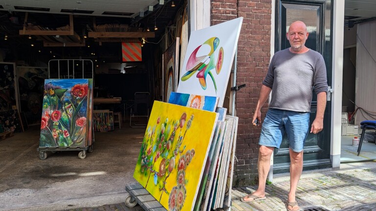 Man in casual kleding staat buiten met verschillende kleurrijke schilderijen tegen een bakstenen muur.