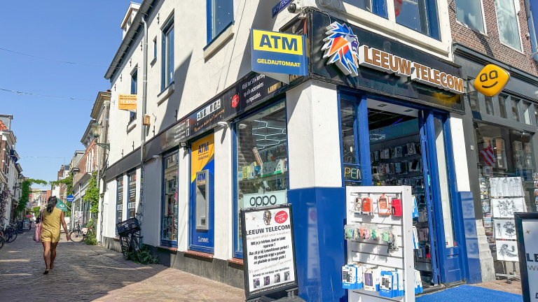 Straatbeeld met een winkel genaamd Leeuw Telecom, met telefoonaccessoires en een geldautomaat naast de ingang.