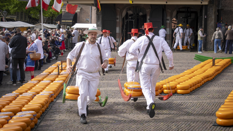 Mannen in witte pakken dragen kazen op dragers tijdens een traditionele kaasmarkt met toeschouwers.