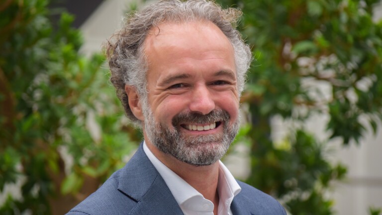 Lachende man met baard en krullend haar draagt een blauw colbert, met groene planten op de achtergrond.