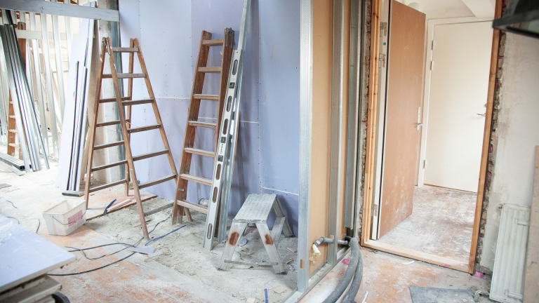 Binnenruimte in verbouwing met ladders, waterpas en bouwafval op de vloer naast een open deur en gipswand