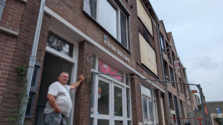 Man staat bij voordeur van bakstenen pand met gevelbordje Infinite skin care en raamtekst TapParfum, achter bouwhek