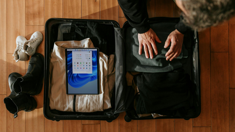 Van bovenaf: persoon pakt kleding in een open koffer op houten vloer, met tablet erop en schoenen ernaast