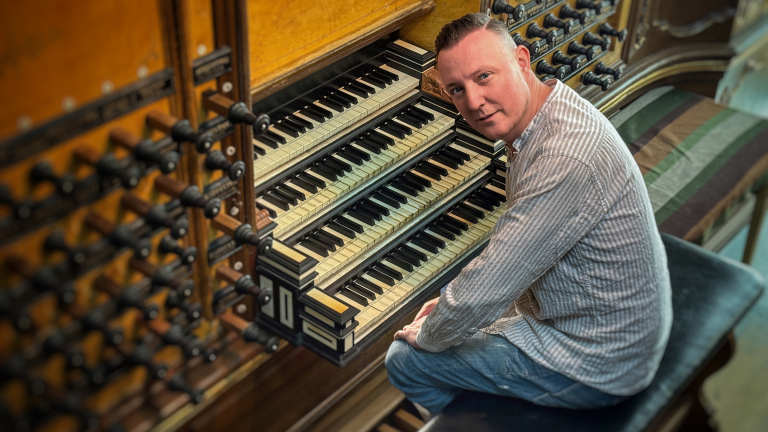 Man zit bij een orgel met meerdere klavieren en registerknoppen en kijkt naar de camera