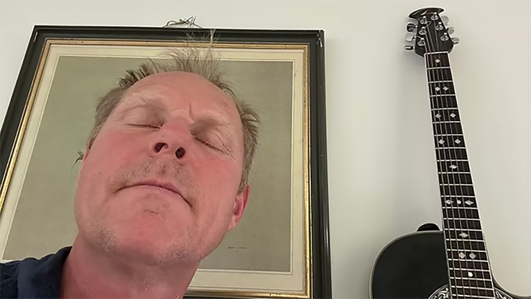 Close-up van een man met gesloten ogen, met een ingelijste afbeelding achter hem en een gitaar rechts in beeld