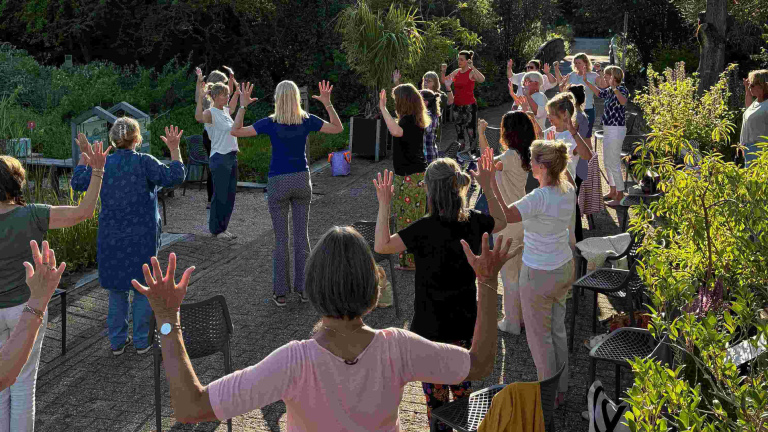 Groep mensen doet buiten een gezamenlijke workout met opgeheven armen op een zonnig terras met groen eromheen