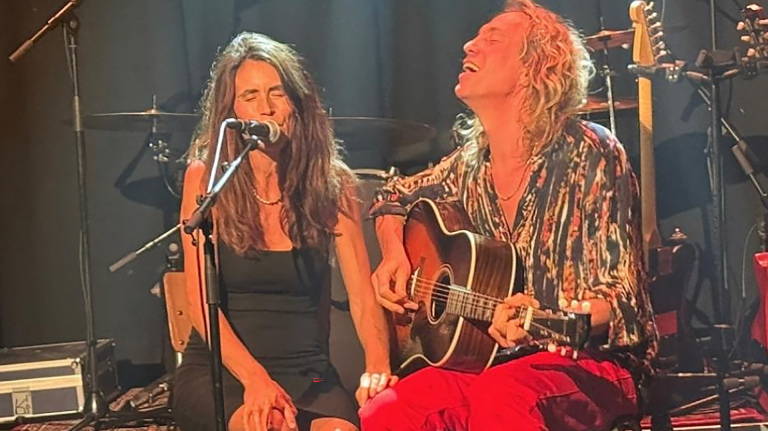 Twee muzikanten op het podium: vrouw zingt in microfoon terwijl man met lang haar akoestische gitaar speelt en meezingt