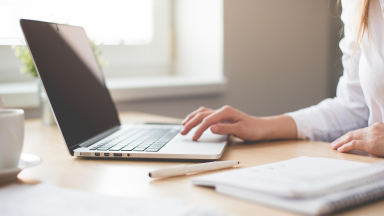 Hand van persoon op laptop op een bureau met notitieboek en kop koffie in een lichte werkomgeving