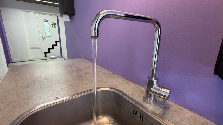 Chromen keukenkraan met stromend water boven een roestvrijstalen gootsteen, tegen een paarse muur