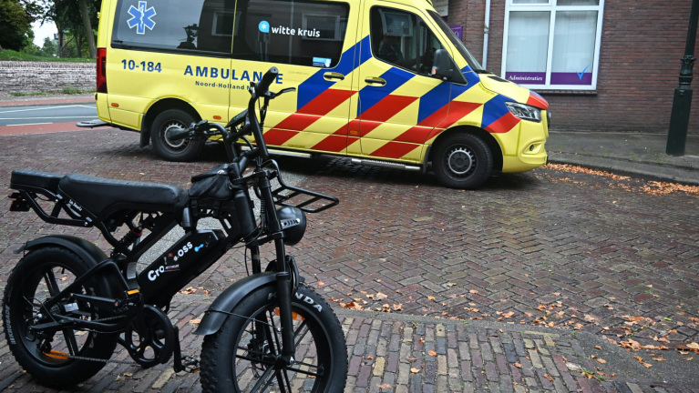 Zwarte elektrische fiets op bakstenen straat, met gele ambulance van Witte Kruis op de achtergrond