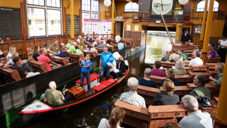 Publiek zit langs een binnenkanaal terwijl een rondvaartboot met gidsen en ballonnen door een hal met grote klok vaart