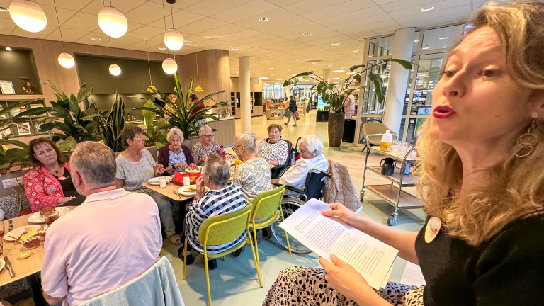 Vrouw leest voor uit papier terwijl een groep ouderen rond een tafel koffie drinkt in een lichte ontmoetingsruimte