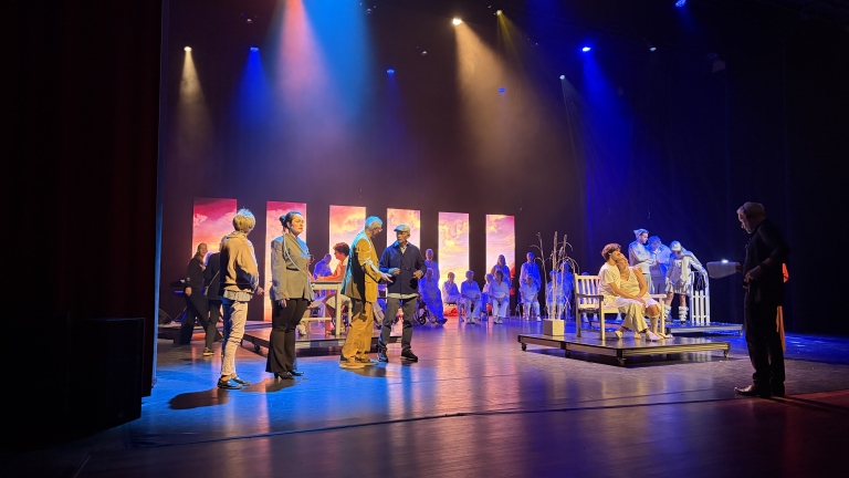 Toneelrepetitie met acteurs onder kleurrijke spotlights, met achteraan een groep in wit en een bankje op het podium
