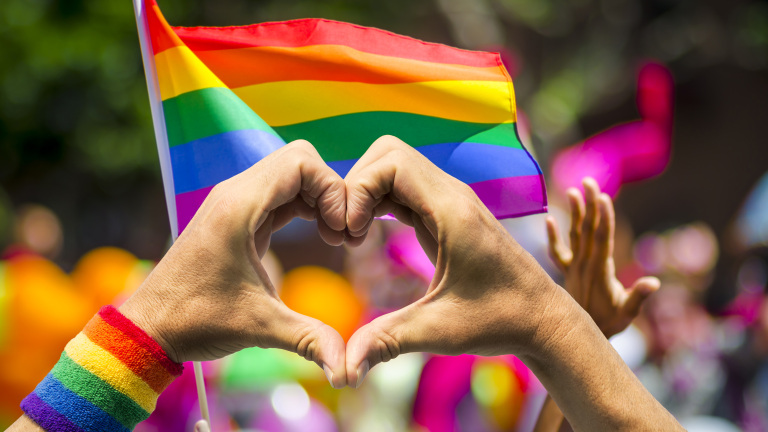 Handen vormen een hartje voor een wapperende regenboogvlag tijdens een Pride-evenement