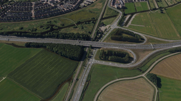 Luchtfoto van snelweg met verkeersknooppunt en viaduct over spoorlijn, omringd door velden, water en woonwijk