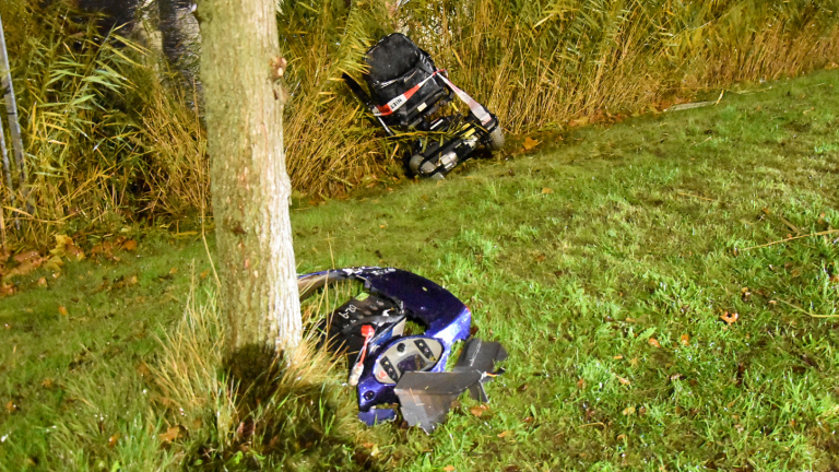 Brandweer en politie zijn aanwezig op een straat met blauwe lichten van een brandweerwagen die de omgeving verlichten, 's nachts geparkeerde auto's langs de weg en een showroom op de achtergrond.