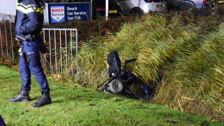 Brandweer en politie zijn aanwezig op een straat met blauwe lichten van een brandweerwagen die de omgeving verlichten, 's nachts geparkeerde auto's langs de weg en een showroom op de achtergrond.