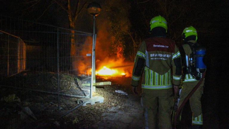 Brandweermannen in uniform bestrijden 's nachts een felle brand bij een omheind gebied.