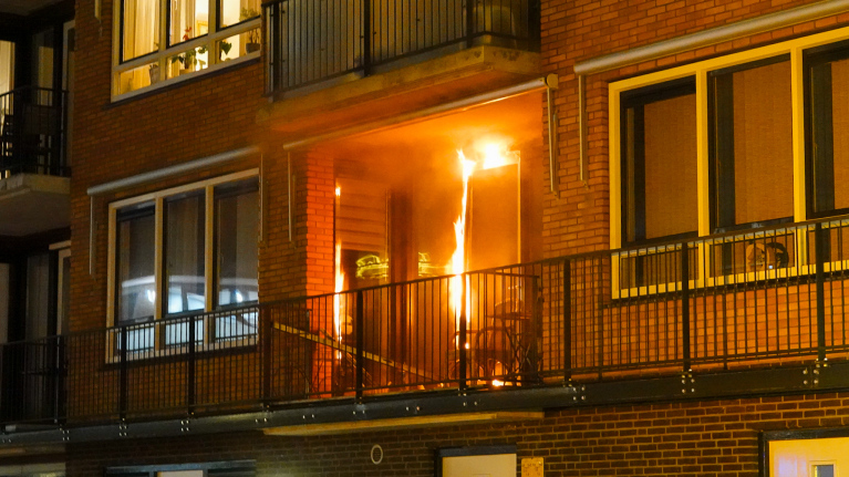 Brand op een balkon van een appartementencomplex, met vlammen zichtbaar rond de deur, naast gesloten ramen.