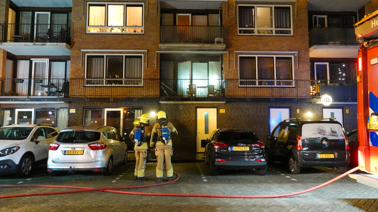 Brand op een balkon van een appartementencomplex, met vlammen zichtbaar rond de deur, naast gesloten ramen.