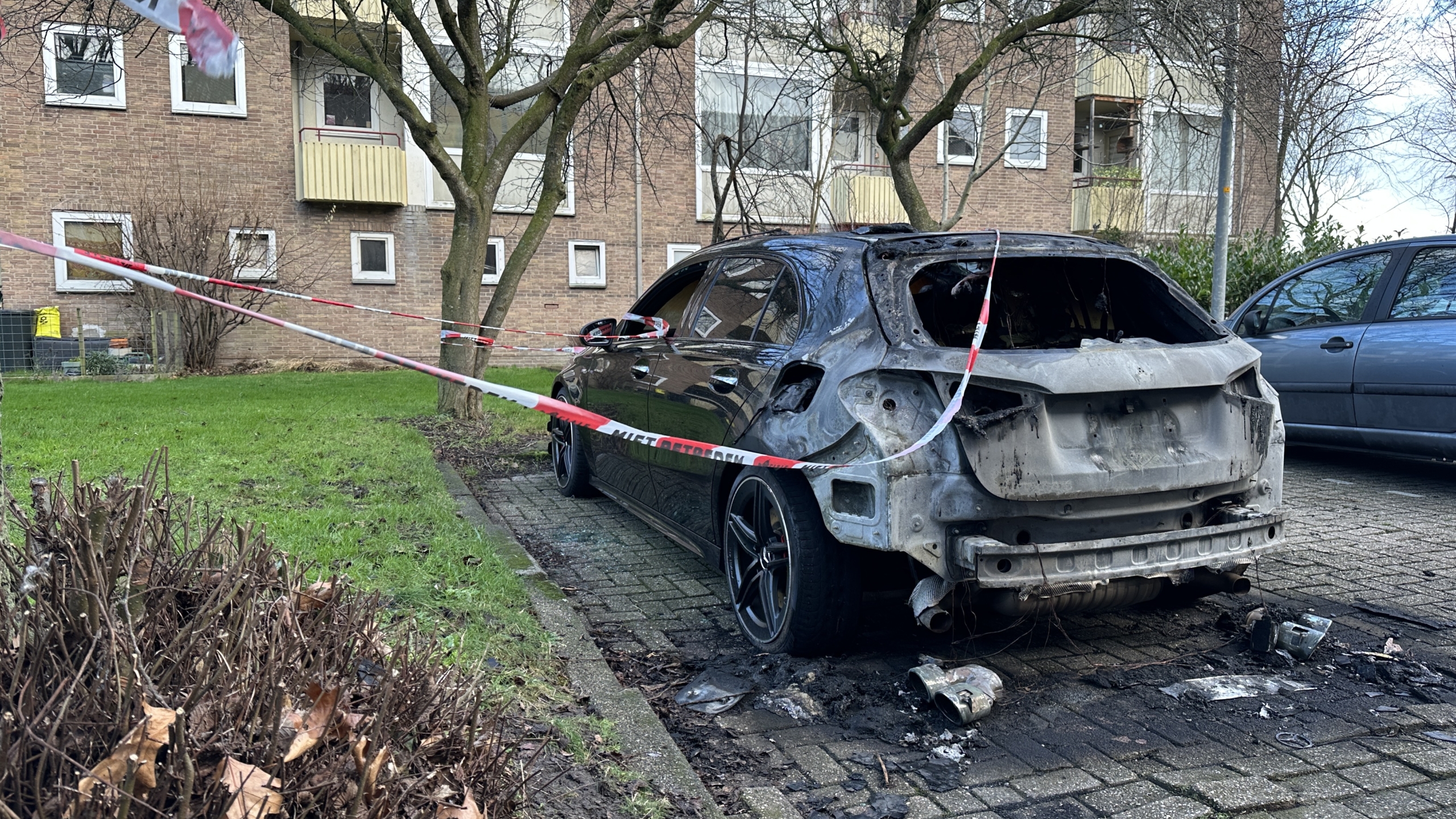 Uitgebrande zwarte auto afgezet met tape waarop "Niet betreden" staat, geparkeerd langs een boom.