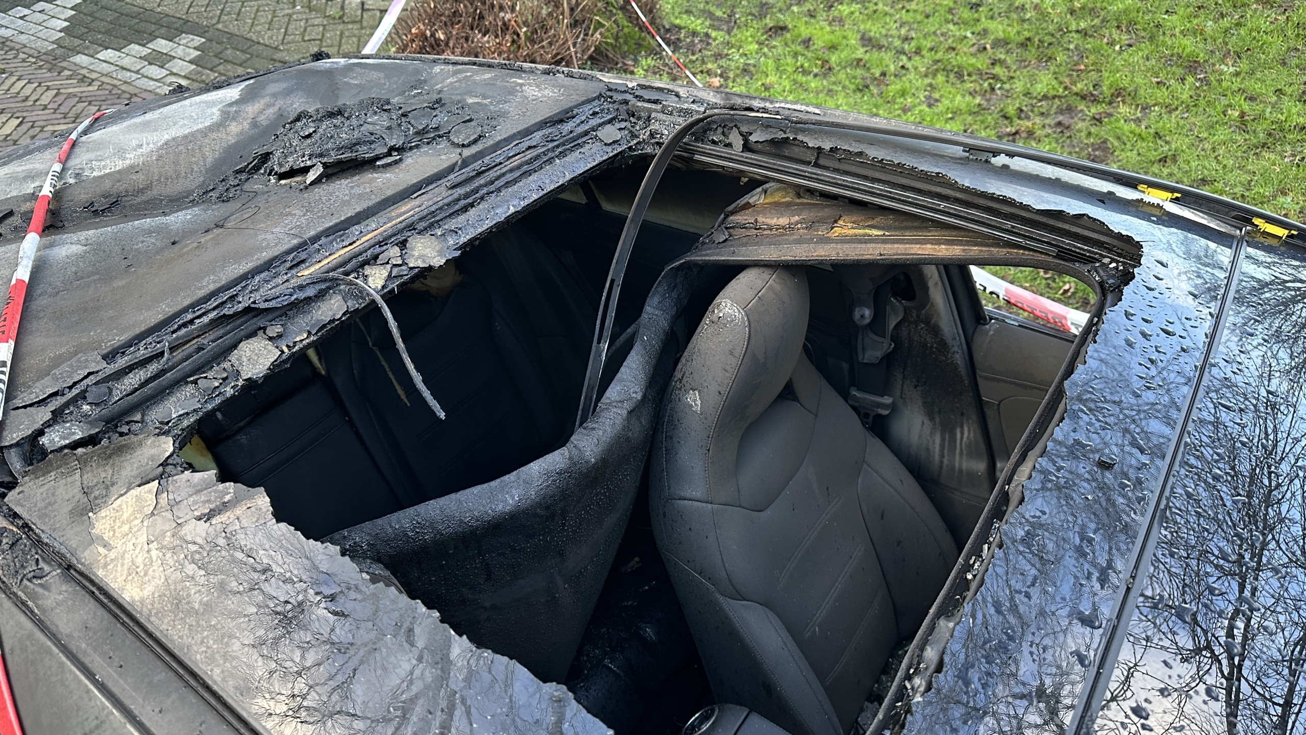 Uitgebrande zwarte auto afgezet met tape waarop "Niet betreden" staat, geparkeerd langs een boom.