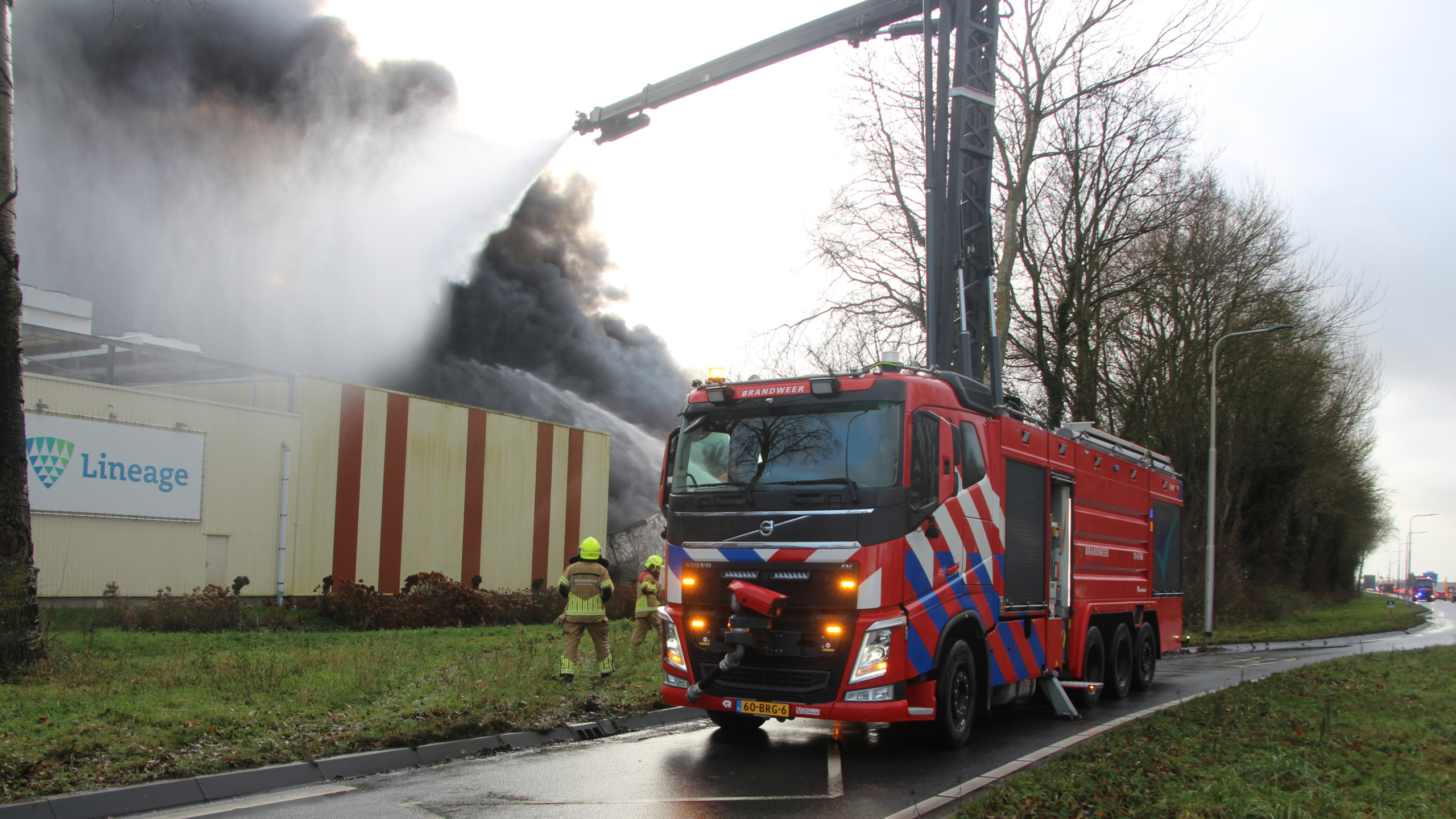 Brandweermannen blussen een intense brand in een loods, waar feloranje vlammen en dikke rookwolken uit het dak slaan.