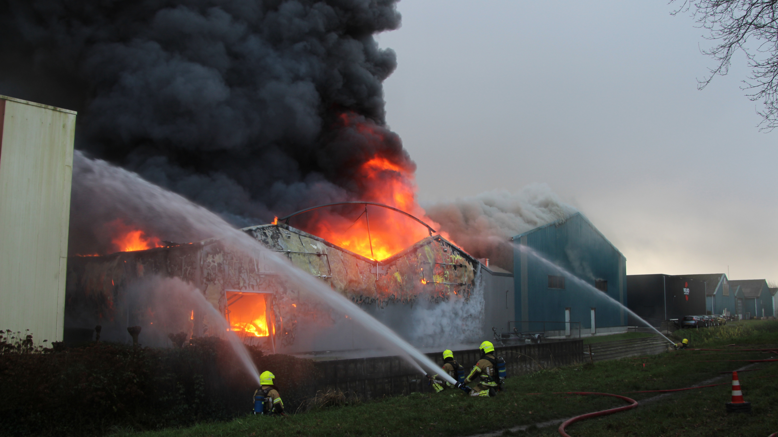 Brandweermannen blussen een intense brand in een loods, waar feloranje vlammen en dikke rookwolken uit het dak slaan.