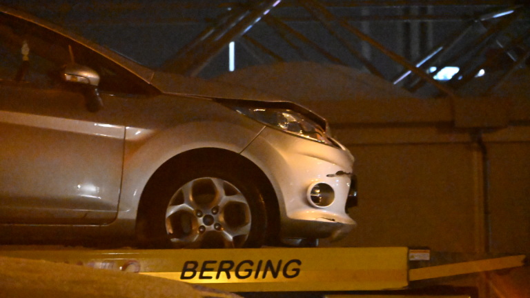 Een auto wordt 's nachts op een takelwagen geladen, begeleid door een persoon in een oranje veiligheidsvest.