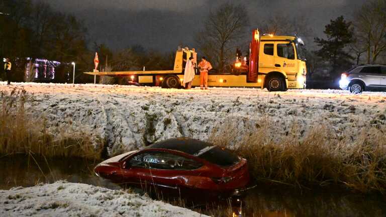 Een rode auto deels ondergedompeld in een sloot langs een besneeuwde weg, met een bergingsvoertuig en mensen erboven.