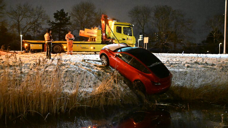 Een rode auto deels ondergedompeld in een sloot langs een besneeuwde weg, met een bergingsvoertuig en mensen erboven.