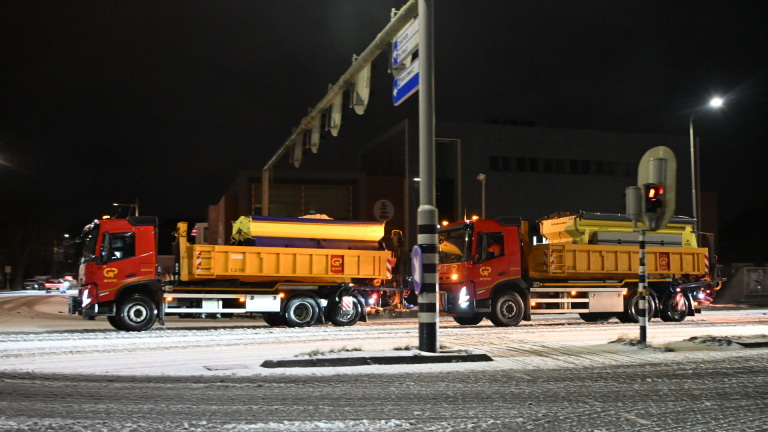 Sneeuwschuiver rijdt 's nachts over besneeuwde weg langs showroom met geparkeerde auto's.