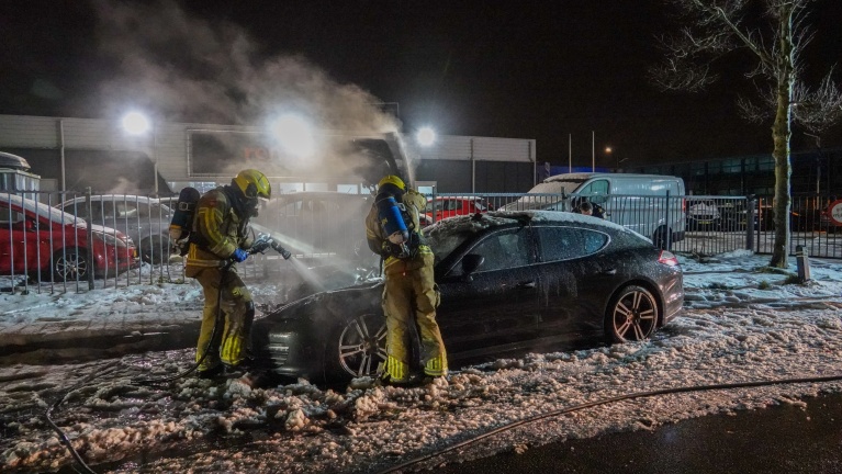 Brandweerlieden blussen 's nachts een brand in een zwarte auto, omringd door schuim en water op de grond.