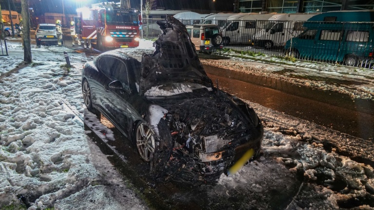 Brandweerlieden blussen 's nachts een brand in een zwarte auto, omringd door schuim en water op de grond.