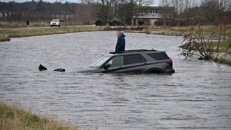 Politie- en brandweerwagens staan aan de kant van de weg bij een half gezonken auto in een sloot, met een agent die toekijkt.