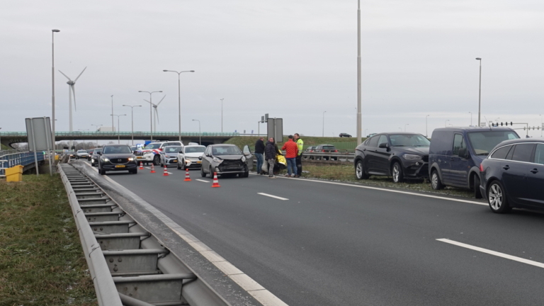 Voorbumperloze zilvergrijze auto op snelweg naast mensen in winterkleding die naast een berm samenkomen, met een verkeerskegel ervoor.